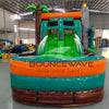 Close-up front view of Dino Dive Dual Lane Commercial Inflatable Combo showing the entrance area. Two realistic T-Rex figures stand on either side of the archway, which is designed to look like a gray stone castle with green accents. A slide is visible on one side, and inflatable vegetation adds to the jungle theme. The "BOUNCEWAVE Inflatable Sales" logo is prominently displayed on the base. Set in a warehouse with various other inflatable products visible in the background.