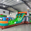 Side view of Dino Dive Dual Lane Commercial Inflatable Combo set up in a large indoor facility. Shows the full length of the inflatable with gray castle structure, green accents, dual slides, and T-Rex figures. Inflatable plants and a splash pool area are visible. Various inflatable products and warehouse materials in the background demonstrate its commercial setting.