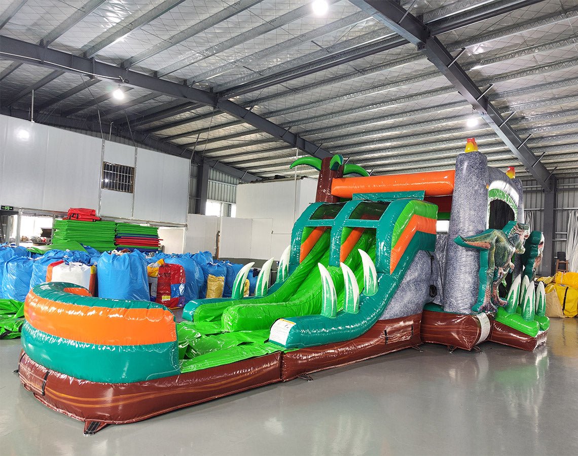 Side view of Dino Dive Dual Lane Commercial Inflatable Combo set up in a large indoor facility. Shows the full length of the inflatable with gray castle structure, green accents, dual slides, and T-Rex figures. Inflatable plants and a splash pool area are visible. Various inflatable products and warehouse materials in the background demonstrate its commercial setting.