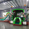 close side view of Dino Dive Dual Lane Commercial Inflatable Combo set up in a spacious warehouse. The inflatable features a gray castle structure with green trim, flame-topped towers, and two imposing T-Rex figures guarding the entrance. Dual slides lead to a splash pool area, with inflatable plants adding to the prehistoric theme. Other warehouse equipment and inflatables are visible in the background.