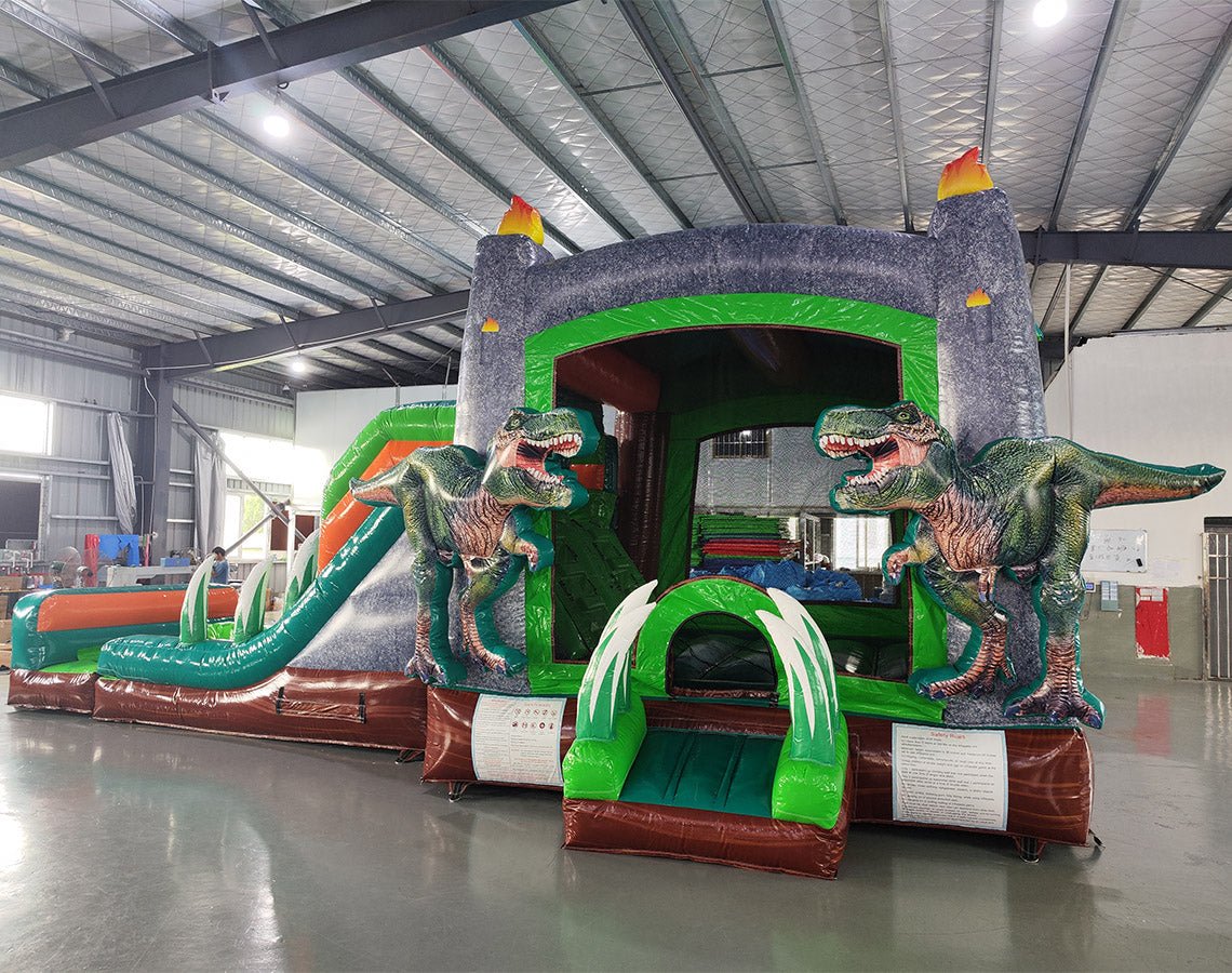 close side view of Dino Dive Dual Lane Commercial Inflatable Combo set up in a spacious warehouse. The inflatable features a gray castle structure with green trim, flame-topped towers, and two imposing T-Rex figures guarding the entrance. Dual slides lead to a splash pool area, with inflatable plants adding to the prehistoric theme. Other warehouse equipment and inflatables are visible in the background.