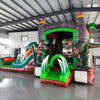 Wide-angle view of the Dino Dive Econo Combo featuring a green and orange inflatable structure with volcano-shaped bounce house, slide, splash pool, and multiple realistic dinosaur figures. Set up in a spacious indoor warehouse with metal roofing and other inflatable products visible in the background.