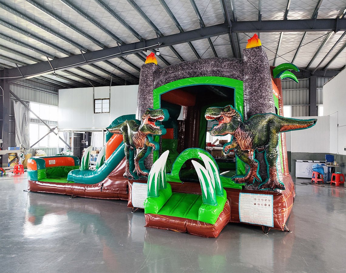 Wide-angle view of the Dino Dive Econo Combo featuring a green and orange inflatable structure with volcano-shaped bounce house, slide, splash pool, and multiple realistic dinosaur figures. Set up in a spacious indoor warehouse with metal roofing and other inflatable products visible in the background.