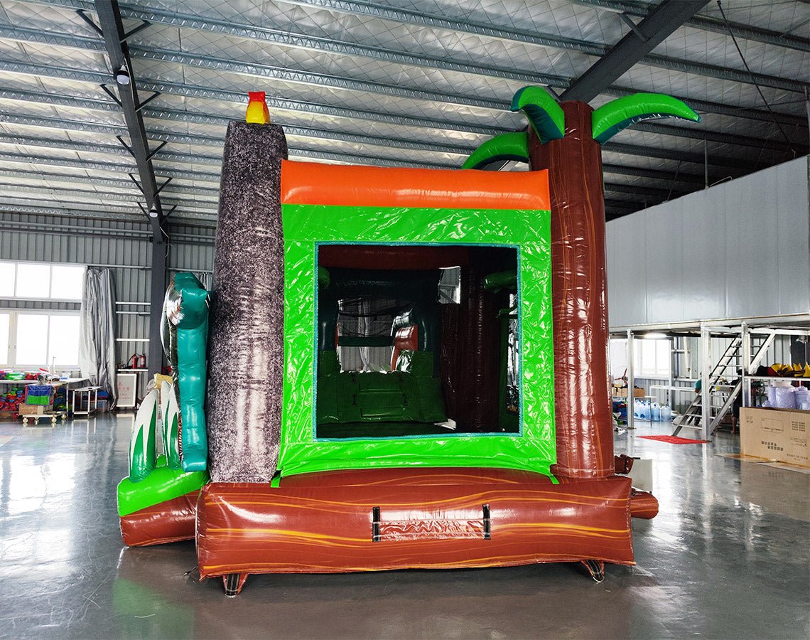 Rear view of Dino Dive Econo Combo highlighting the green bounce house structure with volcano design, orange slide, and splash pool area. Set up in an indoor warehouse with metal ceiling and other inflatable products visible.