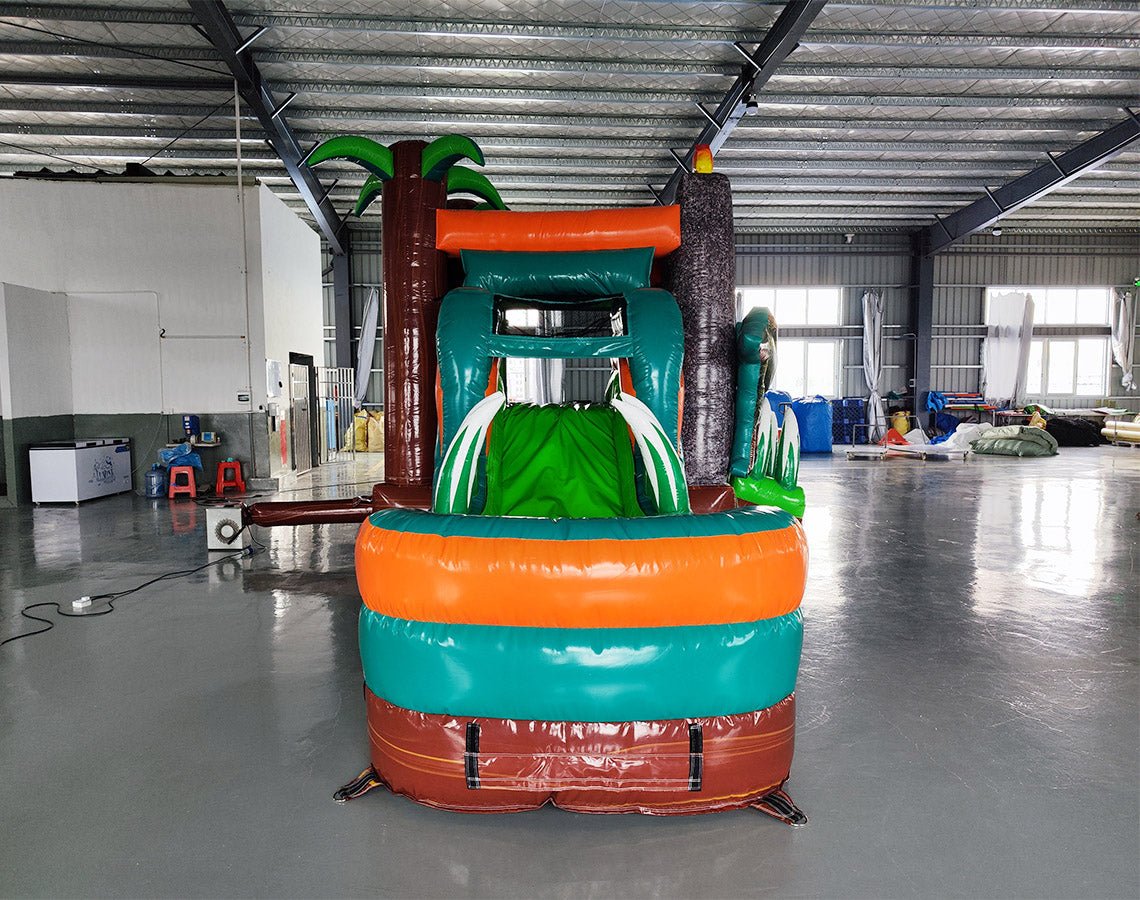 Large inflatable dinosaur-themed combo featuring a bounce house with volcano design, attached slide, and splash pool. The structure has green, orange, and brown colors with realistic dinosaur figures guarding the entrance. Set up in an indoor warehouse with metal roofing and other equipment visible in the background.