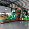 Front view of Dino Dive Econo Combo, showcasing the bounce house entrance with volcano design, green slide, and orange splash pool area. Large, lifelike dinosaur figures flank the entrance. Displayed in an indoor warehouse with various equipment and inflatables visible.