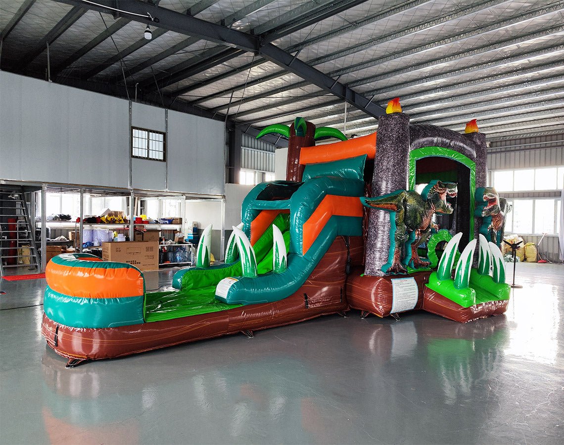 Front view of Dino Dive Econo Combo, showcasing the bounce house entrance with volcano design, green slide, and orange splash pool area. Large, lifelike dinosaur figures flank the entrance. Displayed in an indoor warehouse with various equipment and inflatables visible.