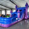 Side view of Enchanted Castle Dual Lane Commercial Inflatable Combo set up in an indoor facility. The inflatable structure shows its full length, featuring blue walls, purple towers with blue conical tops, dual slides, and a large splash pool area. The unicorn-themed banner and colorful front panel are visible from this angle. The warehouse setting with metal roofing, support beams, and other inflatable products provides scale context.