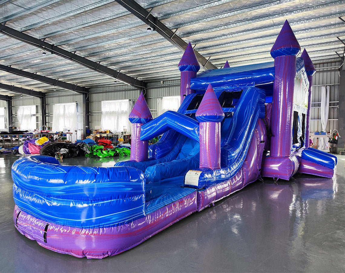 Side view of Enchanted Castle Dual Lane Commercial Inflatable Combo set up in an indoor facility. The inflatable structure shows its full length, featuring blue walls, purple towers with blue conical tops, dual slides, and a large splash pool area. The unicorn-themed banner and colorful front panel are visible from this angle. The warehouse setting with metal roofing, support beams, and other inflatable products provides scale context.