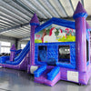 Wide-angle view of Enchanted Castle Dual Lane Commercial Inflatable Combo in a spacious warehouse. The inflatable features blue walls, purple towers with blue tops, dual slides, and a large splash pool area. A unicorn-themed banner stretches across the top, and the front panel displays a colorful scene with unicorns and a castle. Various other inflatable products and warehouse equipment are visible, showcasing its size and commercial setting.