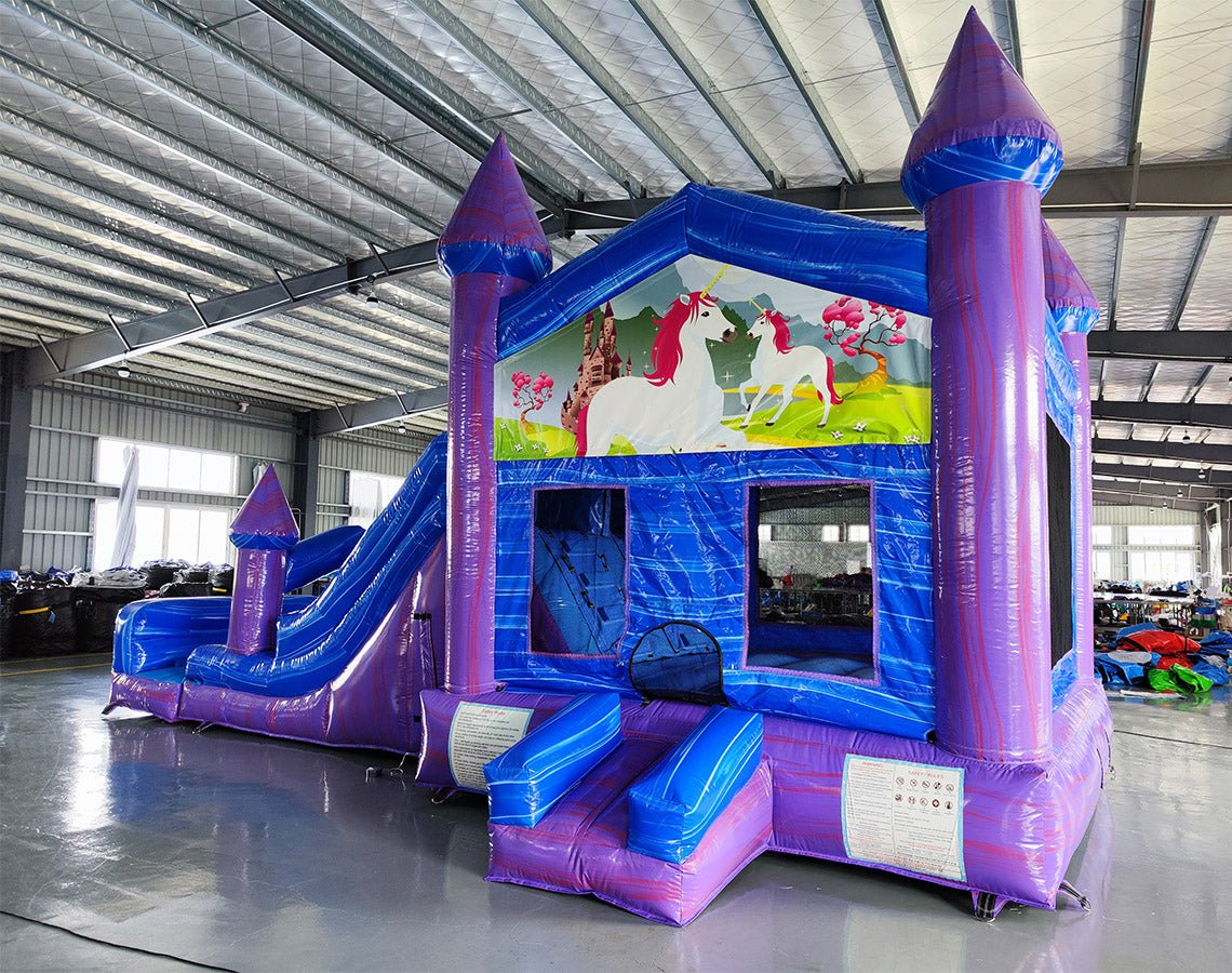 Wide-angle view of Enchanted Castle Dual Lane Commercial Inflatable Combo in a spacious warehouse. The inflatable features blue walls, purple towers with blue tops, dual slides, and a large splash pool area. A unicorn-themed banner stretches across the top, and the front panel displays a colorful scene with unicorns and a castle. Various other inflatable products and warehouse equipment are visible, showcasing its size and commercial setting.