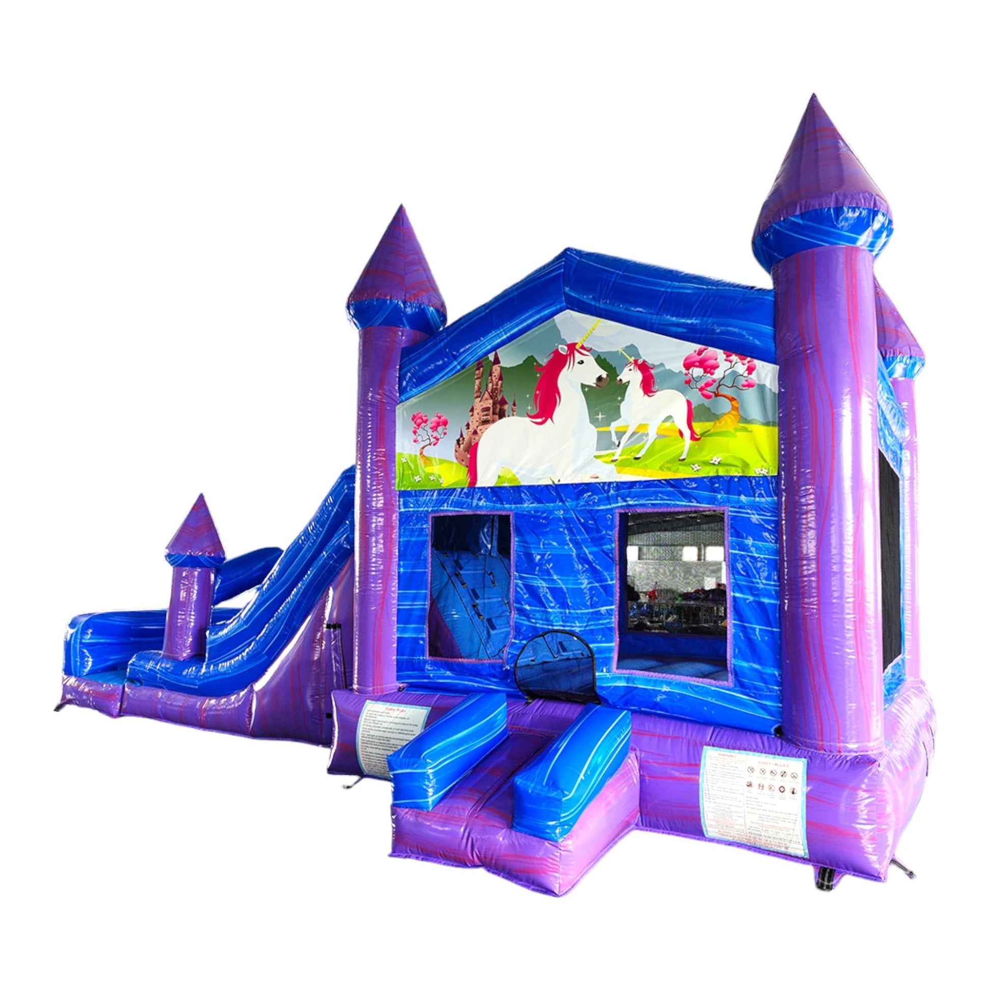 Side view of Enchanted Castle Dual Lane Commercial Inflatable Combo featuring a vibrant blue and purple color scheme. The structure has tall purple towers with blue conical tops, dual slides, and a unicorn-themed banner across the top. A colorful scene with unicorns and a castle is displayed on the front panel. Set up in a large indoor warehouse with metal roofing and other inflatable products visible in the background.