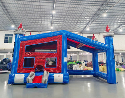 Front view of Euro Baja Commercial Grade Canopy Bounce House in a warehouse setting. Features a blue inflatable structure with red interior walls, mesh windows, and four tall blue pillars topped with white and red castle-like turrets. The entrance has a step and safety instructions visible. Various inflatable products and warehouse equipment are seen in the background.