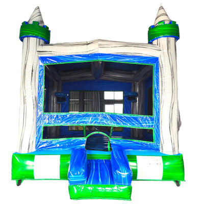 Front view of Euro Gray Commercial Grade Bounce House featuring a white inflatable castle structure with gray stone-like texture. The bounce house has two tall turrets with green conical tops and blue flags. The entrance is framed by blue inflatable pillars and a green archway. A blue and green slide is attached to the right side. The structure is set up in a large indoor warehouse with metal roofing and other inflatable products visible in the background.