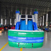 Front view of Euro Green Castle Dual Lane Commercial Inflatable Combo set up in an indoor facility. The inflatable castle structure has white towers with blue turrets and green conical tops. It features dual blue slides, a bounce house entrance with mesh windows, and a circular splash pool. Various other inflatable products and warehouse equipment are visible in the background, demonstrating its size and commercial setting.