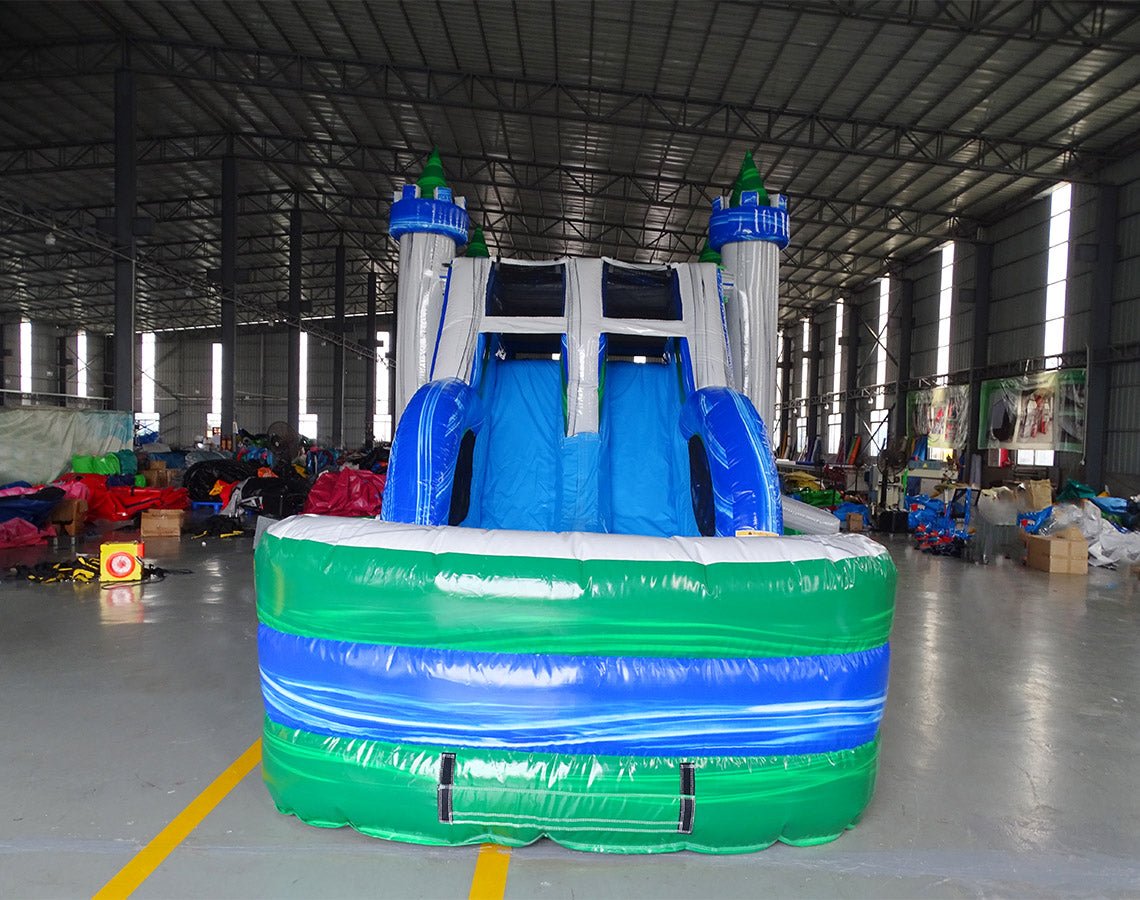 Front view of Euro Green Castle Dual Lane Commercial Inflatable Combo set up in an indoor facility. The inflatable castle structure has white towers with blue turrets and green conical tops. It features dual blue slides, a bounce house entrance with mesh windows, and a circular splash pool. Various other inflatable products and warehouse equipment are visible in the background, demonstrating its size and commercial setting.