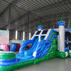 Large inflatable Euro Green Castle Dual Lane Commercial Combo featuring a castle-themed design with white towers, blue turrets, and green conical tops. The structure includes dual blue slides, a bounce house area with mesh windows, and a circular splash pool. Set up in a spacious warehouse with metal roofing and other inflatable products visible in the background.