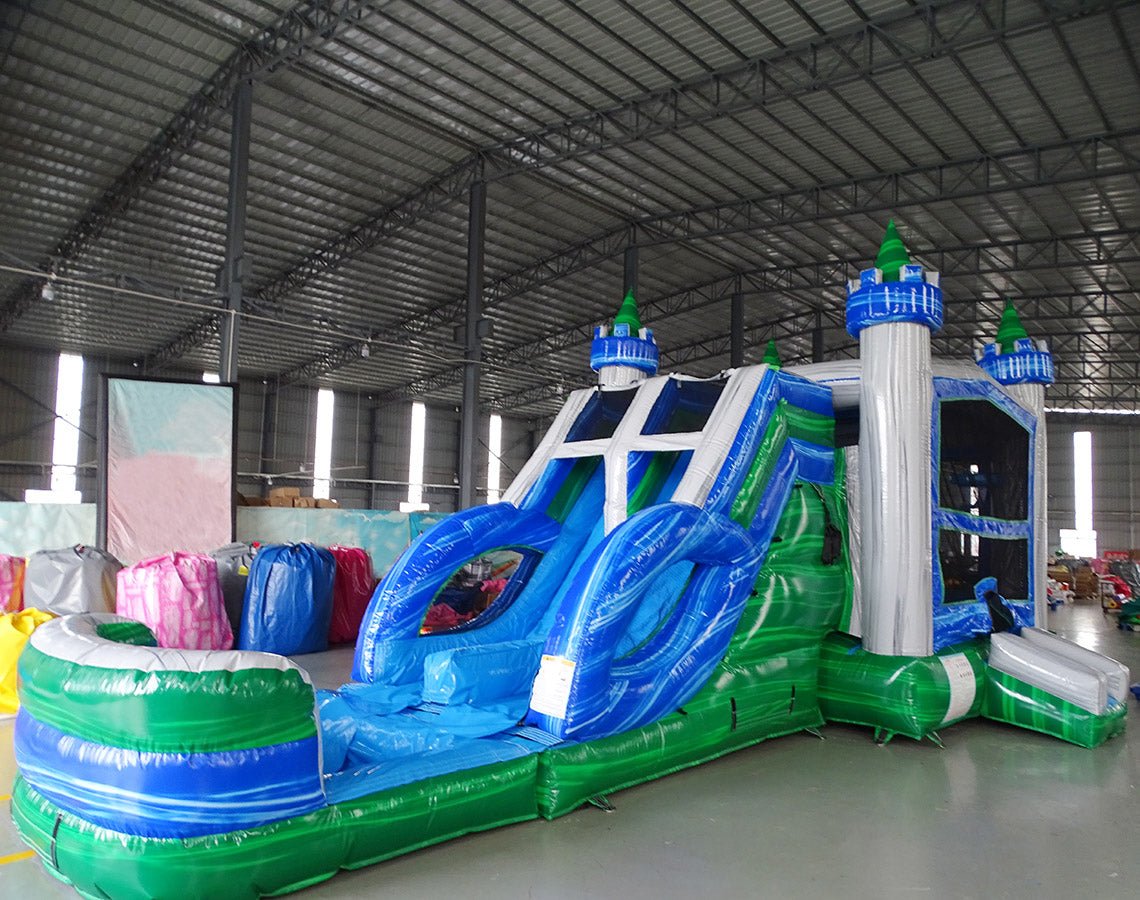 Large inflatable Euro Green Castle Dual Lane Commercial Combo featuring a castle-themed design with white towers, blue turrets, and green conical tops. The structure includes dual blue slides, a bounce house area with mesh windows, and a circular splash pool. Set up in a spacious warehouse with metal roofing and other inflatable products visible in the background.