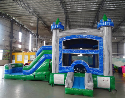 Wide-angle view of Euro Green Gush Commercial Econo Inflatable Wet/Dry Combo featuring a blue and silver castle-themed bounce house with green turrets, slide, and splash pool area. The inflatable is set up in a large indoor warehouse with metal roofing, bright lighting, and other inflatable products visible.