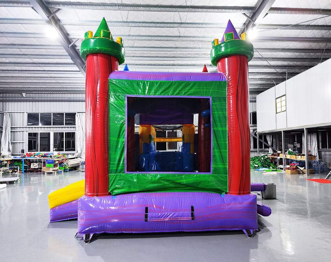 Colorful inflatable castle-themed combo featuring a bounce house with red turrets, green roof, and purple base. Includes a yellow slide and multicolored splash pool. Set up in an indoor warehouse with metal roofing visible.