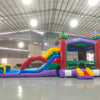 Wide-angle view of Euro Marble Dual Lane Commercial Inflatable Combo (Wet/Dry) in a large indoor facility. Highlights the castle-themed bounce house with colorful turrets, dual curved slides in red and blue, and splash pool area. The structure has vibrant purple, yellow, green, and red colors. Warehouse interior visible with metal roofing, walls, and other inflatable products in the background, providing scale and context for the combo's size.