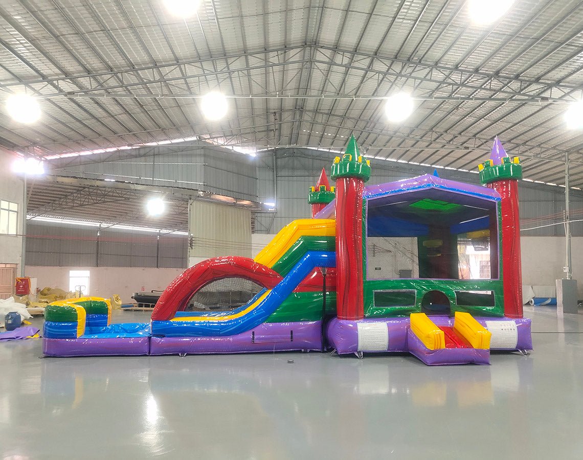 Wide-angle view of Euro Marble Dual Lane Commercial Inflatable Combo (Wet/Dry) in a large indoor facility. Highlights the castle-themed bounce house with colorful turrets, dual curved slides in red and blue, and splash pool area. The structure has vibrant purple, yellow, green, and red colors. Warehouse interior visible with metal roofing, walls, and other inflatable products in the background, providing scale and context for the combo's size.