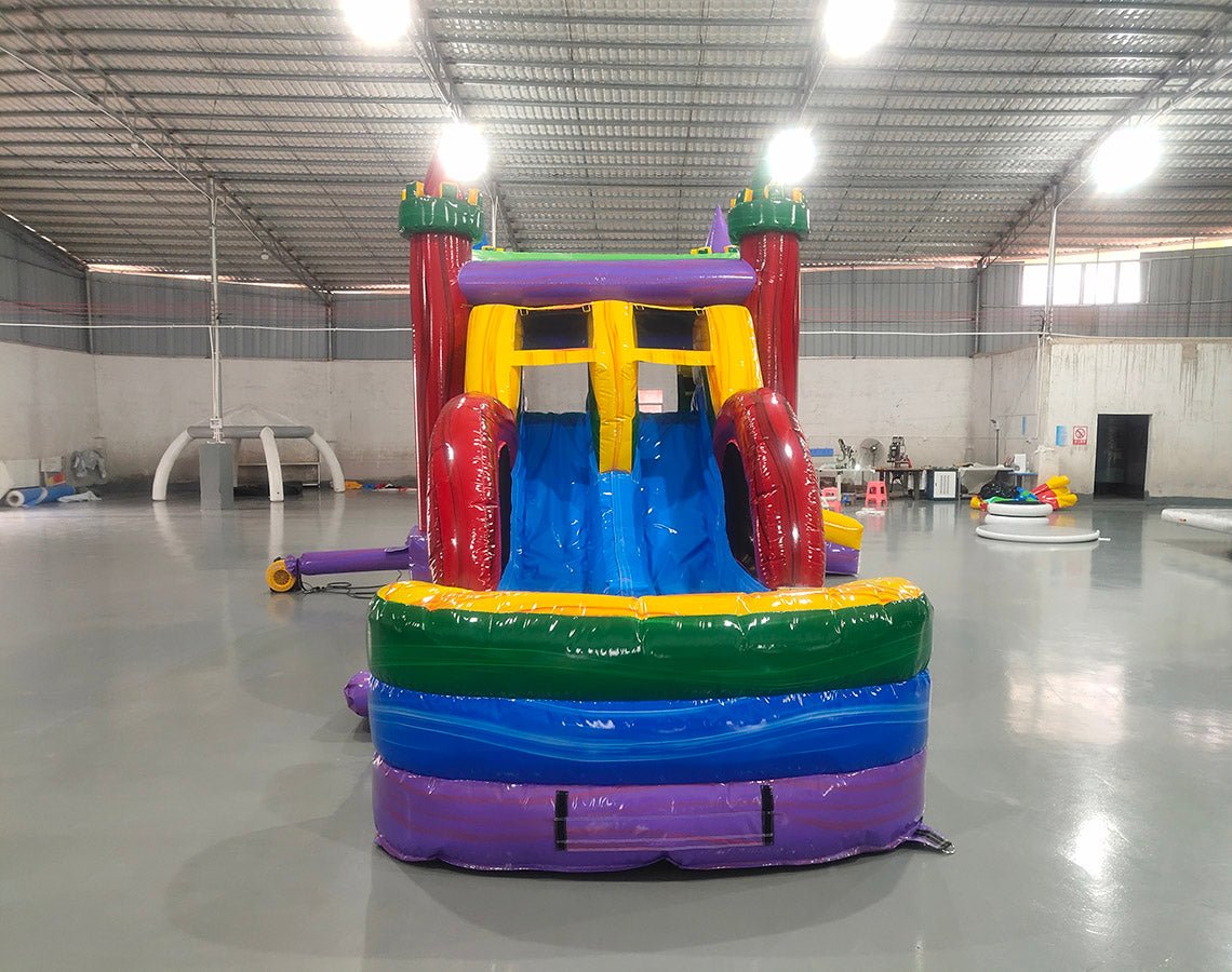 Front view of Euro Marble Dual Lane Commercial Inflatable Combo (Wet/Dry) set up in a large indoor warehouse. Features a castle-themed bounce house with colorful turrets, dual curved slides in red and blue, and a splash pool area. The structure has vibrant purple, yellow, green, and red colors. Metal roofing, concrete flooring, and other warehouse equipment visible in the background.