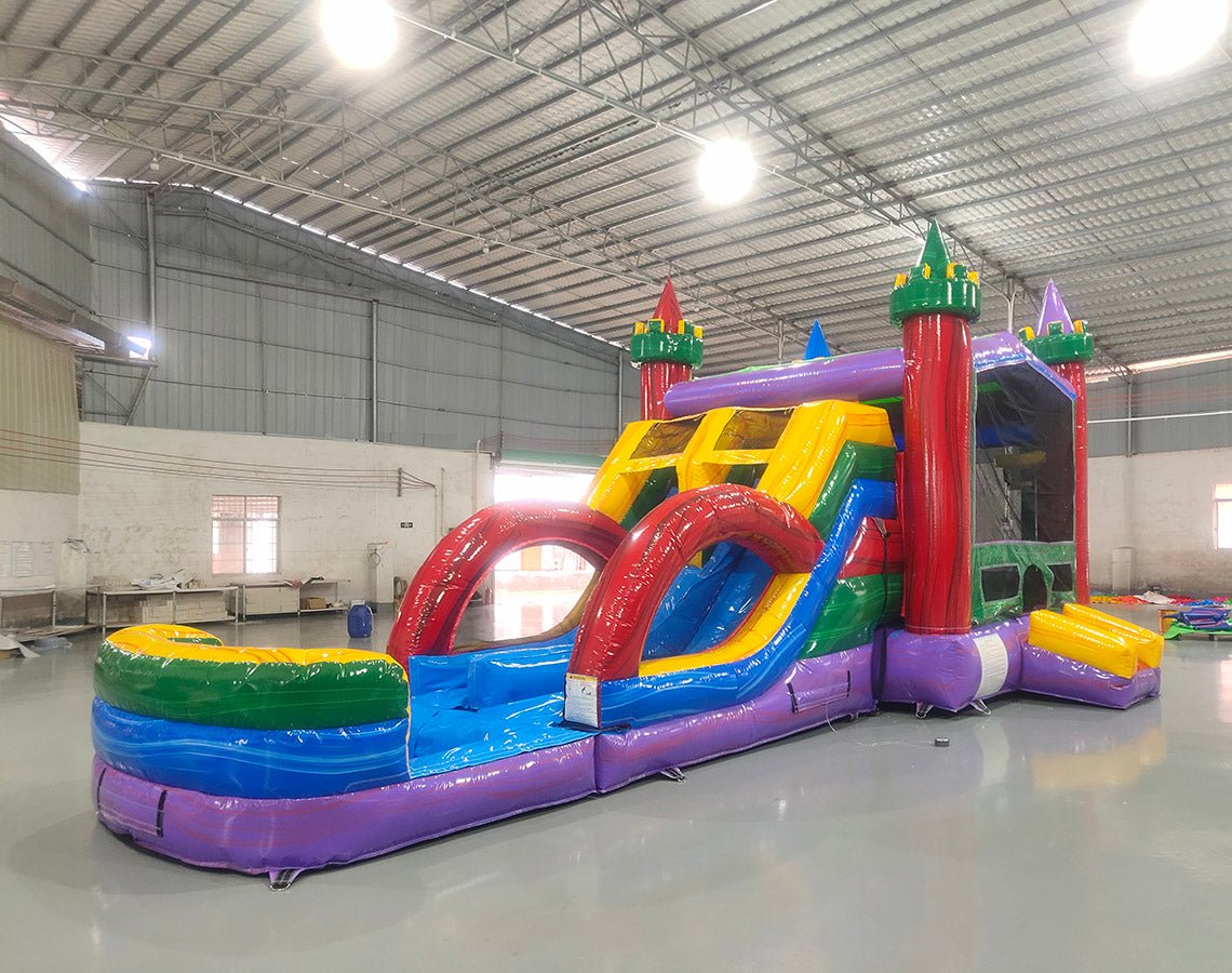 Side view of Euro Marble Dual Lane Commercial Inflatable Combo (Wet/Dry) in a spacious warehouse setting. Showcases the full structure including castle-themed bounce house, dual curved slides in red and blue, and splash pool area. The inflatable features vibrant purple, yellow, green, and red colors. Other inflatable products and warehouse equipment visible, demonstrating its scale in a commercial environment.