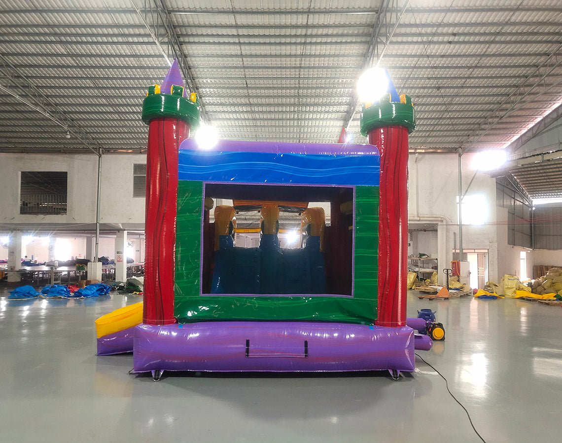 Wide-angle view of Euro Marble Dual Lane Commercial Inflatable Combo (Wet/Dry) set up in a large indoor warehouse. Features a colorful castle-themed structure with red turrets, green battlements, and yellow accents. Two curved slides, one red and one blue, extend from the main structure. The combo includes a bounce house area with mesh windows and a multicolored splash pool at the base. The warehouse setting shows metal roofing, concrete flooring, and other inflatable products in the background.