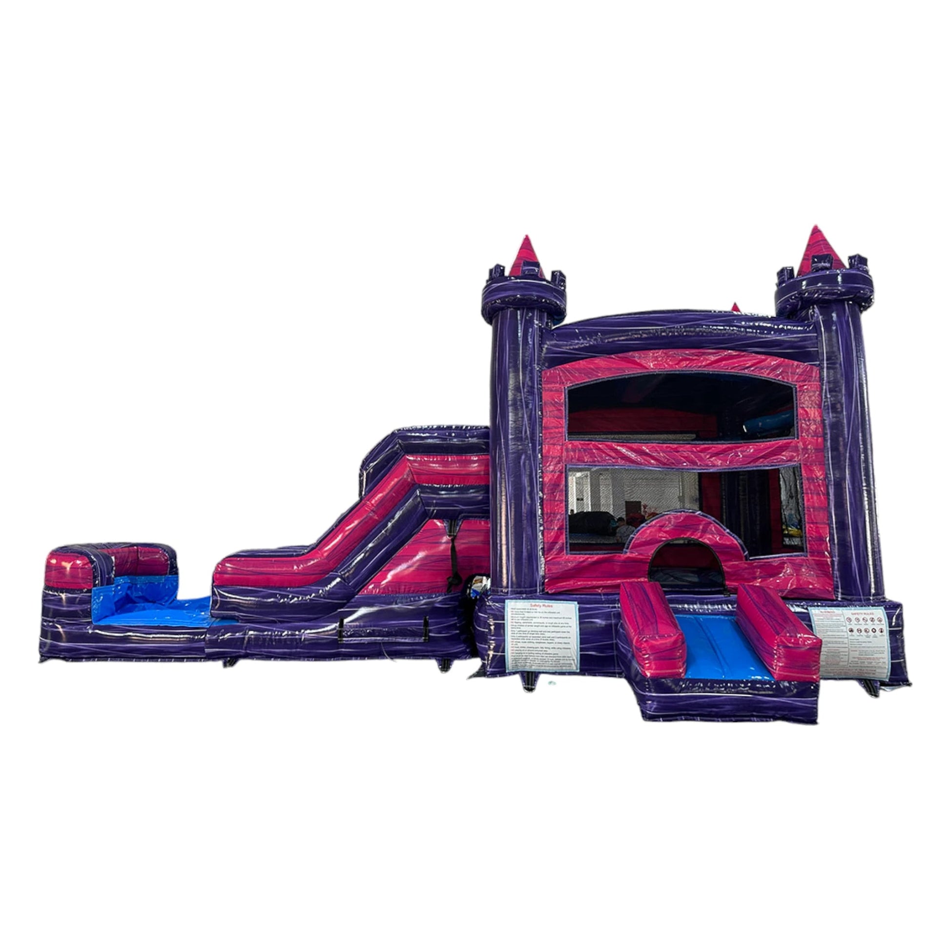 Large inflatable castle-themed combo featuring a bounce house with purple walls, pink turrets, and multicolored accents. Includes a yellow slide and colorful splash pool area. Set up in an indoor warehouse with metal roofing and other equipment visible in the background.