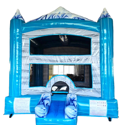 Front view of Everest Falls Commercial Grade Bounce House featuring a large blue and white inflatable structure with a mountain and waterfall theme. The bounce house has two tall blue turrets with red conical tops, a central white peak resembling a snow-capped mountain, and a blue curved slide on the right side. The entrance is framed by inflatable rock-like formations and has a blue step. Set up in a spacious indoor warehouse with metal roofing and fluorescent lighting visible. Other inflatable products an