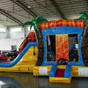 side view of Fiesta Fire Dual Lane Commercial Inflatable Combo set up in a large indoor facility. Highlights the dual orange slides, green palm trees, yellow and blue base, and flame-like decorations. The bounce house entrance and splash pool are prominently displayed. Various other inflatable products and warehouse equipment visible in the background.