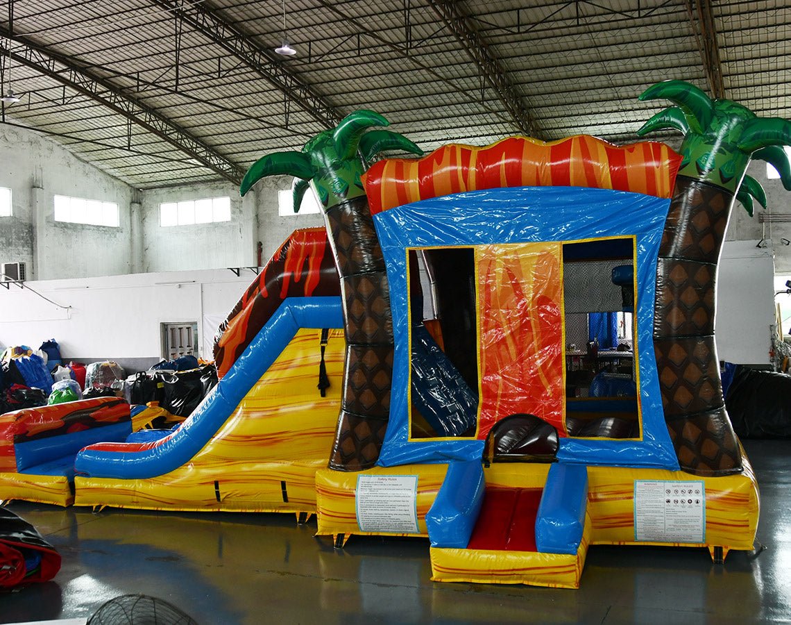 side view of Fiesta Fire Dual Lane Commercial Inflatable Combo set up in a large indoor facility. Highlights the dual orange slides, green palm trees, yellow and blue base, and flame-like decorations. The bounce house entrance and splash pool are prominently displayed. Various other inflatable products and warehouse equipment visible in the background.