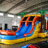 Side angle view of Fiesta Fire Dual Lane Commercial Inflatable Combo in a warehouse environment. Shows the full length of the inflatable with dual orange slides, green palm trees, yellow and blue base, and flame decorations. The bounce house area and splash pool are visible. Other inflatable products and warehouse materials in the background showcase its commercial application.