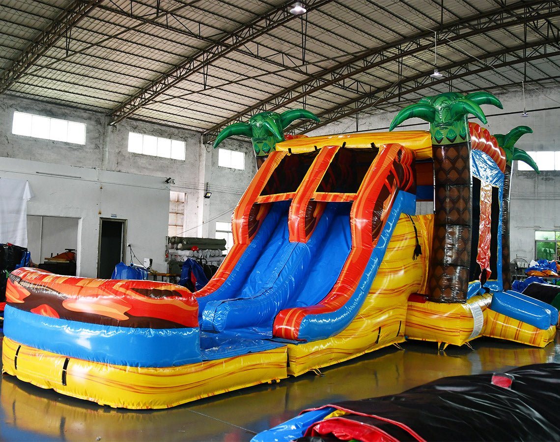 Side angle view of Fiesta Fire Dual Lane Commercial Inflatable Combo in a warehouse environment. Shows the full length of the inflatable with dual orange slides, green palm trees, yellow and blue base, and flame decorations. The bounce house area and splash pool are visible. Other inflatable products and warehouse materials in the background showcase its commercial application.