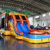 Wide-angle view of Fiesta Fire Dual Lane Commercial Inflatable Combo in a spacious warehouse. The inflatable features dual orange slides, green palm trees, a yellow and blue base, and flame decorations. A large splash pool area is visible. Metal roofing, support beams, and other inflatable products in the background demonstrate its size and commercial setting.