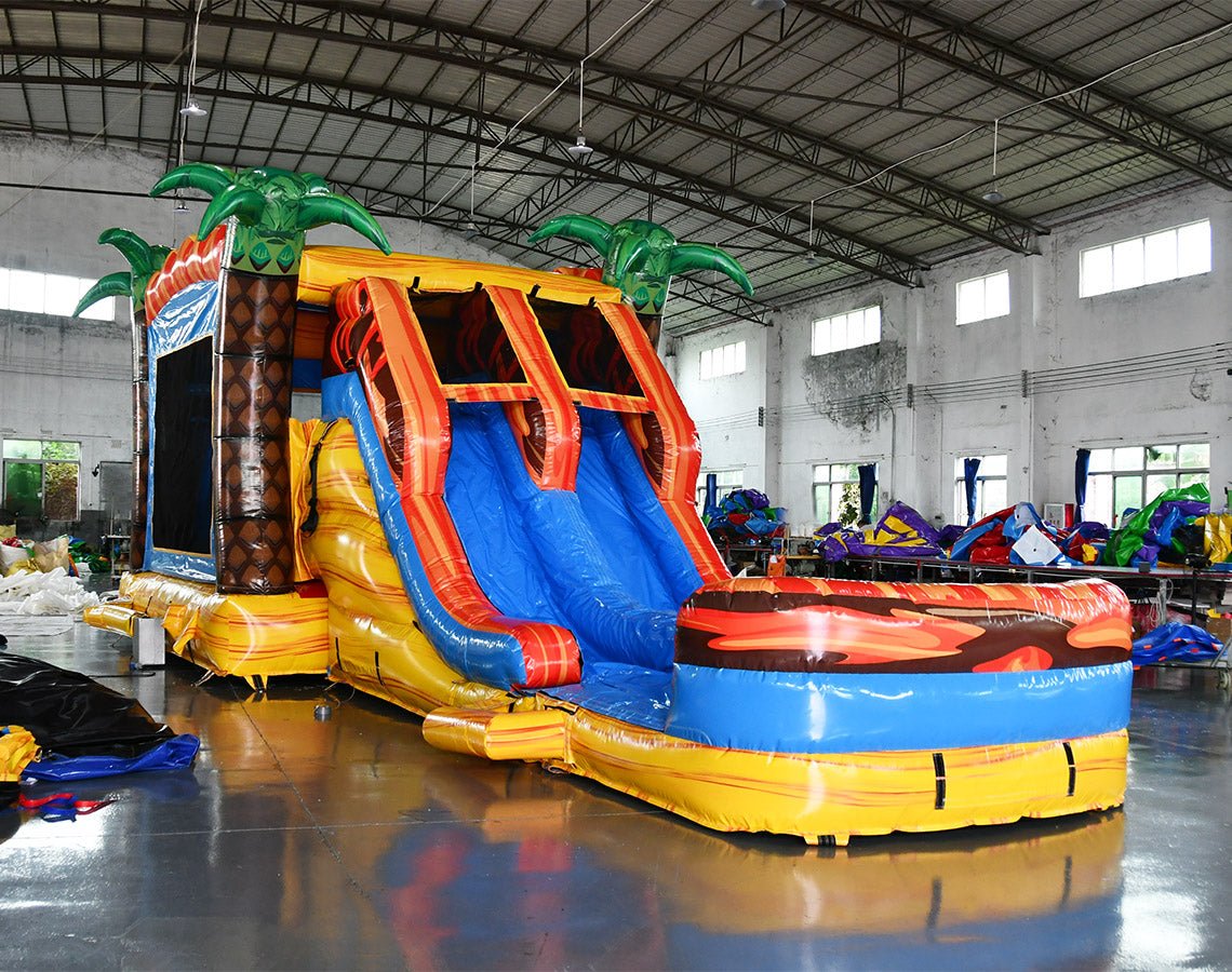 Wide-angle view of Fiesta Fire Dual Lane Commercial Inflatable Combo in a spacious warehouse. The inflatable features dual orange slides, green palm trees, a yellow and blue base, and flame decorations. A large splash pool area is visible. Metal roofing, support beams, and other inflatable products in the background demonstrate its size and commercial setting.