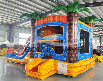 Large inflatable tropical-themed combo featuring a bounce house with palm tree decorations, attached slide, and splash pool. The structure has vibrant orange, red, and blue colors with brown palm tree trunks. Set up in an indoor warehouse with metal roofing visible.