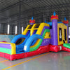 Large inflatable Flat Euro Dual Lane Commercial Combo featuring a castle-themed design with purple towers and yellow flags. The structure includes dual blue slides, a bounce house area with mesh windows, and a circular splash pool. Set up in a spacious warehouse with metal roofing and other inflatable products visible in the background.