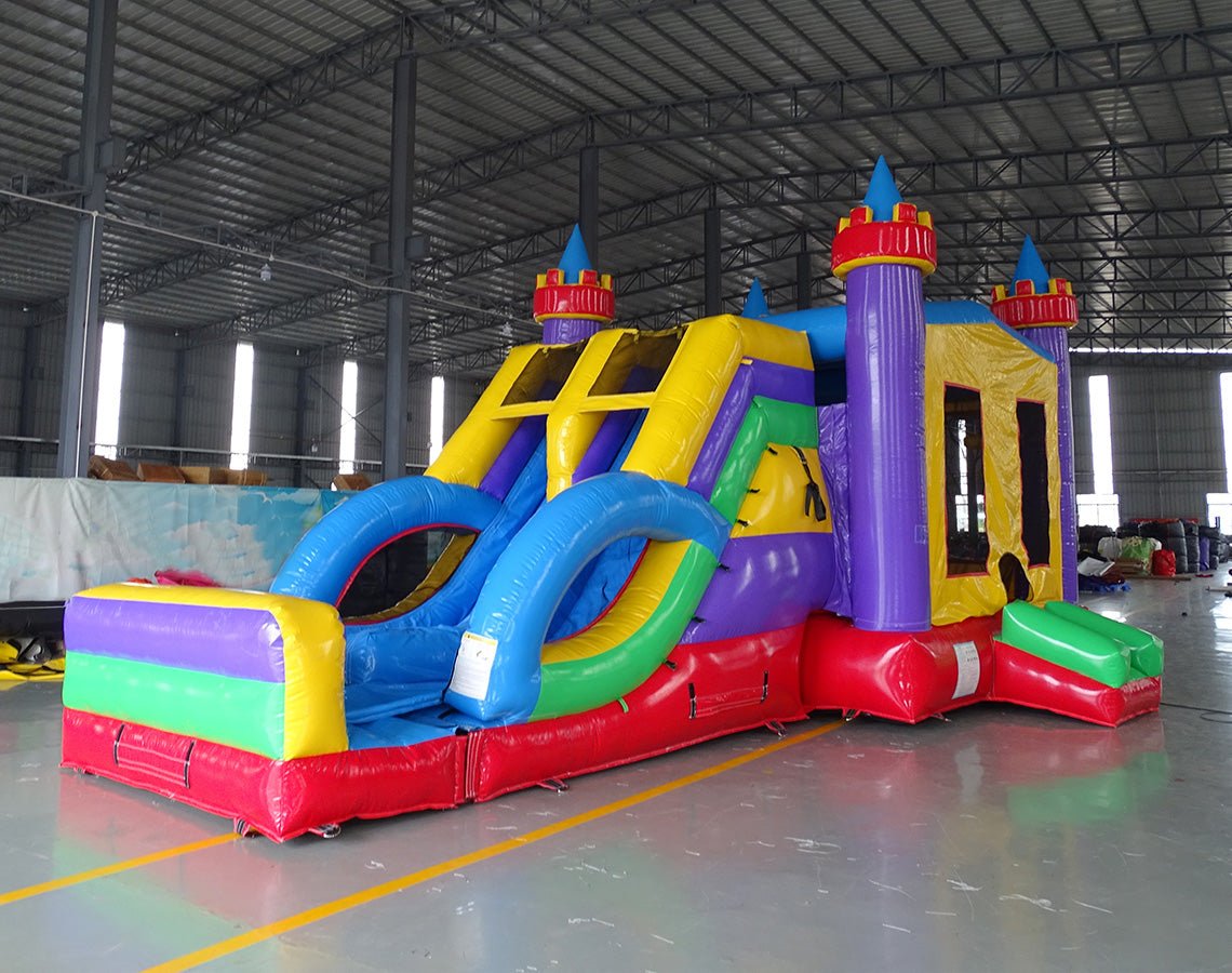 Large inflatable Flat Euro Dual Lane Commercial Combo featuring a castle-themed design with purple towers and yellow flags. The structure includes dual blue slides, a bounce house area with mesh windows, and a circular splash pool. Set up in a spacious warehouse with metal roofing and other inflatable products visible in the background.