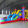 Front view of Flat Euro Dual Lane Commercial Inflatable Combo set up in an indoor facility. The inflatable castle structure has purple towers with yellow flags, dual blue slides, a bounce house entrance with mesh windows, and a circular splash pool. Various other inflatable products and warehouse equipment are visible in the background, demonstrating its size and commercial setting.