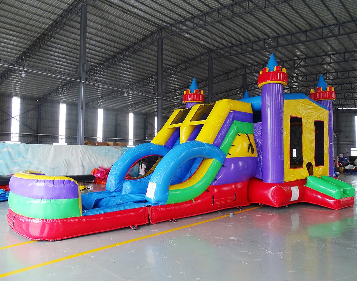 Front view of Flat Euro Dual Lane Commercial Inflatable Combo set up in an indoor facility. The inflatable castle structure has purple towers with yellow flags, dual blue slides, a bounce house entrance with mesh windows, and a circular splash pool. Various other inflatable products and warehouse equipment are visible in the background, demonstrating its size and commercial setting.