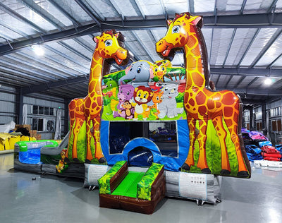 Wide-angle view of the Giraffe Safari Econo Combo in an indoor warehouse. Features two large giraffe inflatables, a bounce house with animal artwork, blue slide, and green obstacles. Other inflatable products and warehouse equipment visible in the background.