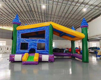 Three-quarter view of Goombay Commercial Grade Canopy Bounce House highlighting its blue inflatable frame, yellow bounce area walls, and yellow canopy top. Four tall blue pillars with white cylindrical turrets and blue conical roofs anchor the corners. Mesh windows allow visibility into the play area. The bounce house is displayed in a large indoor facility with metal roofing, fluorescent lighting, and other inflatable products in the background.