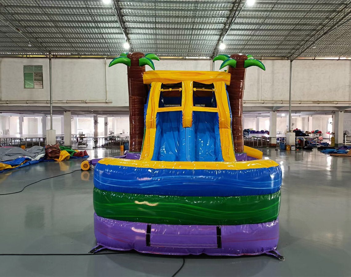 Front view of Goombay Splash Dual Lane Commercial Inflatable Combo set up in a large indoor warehouse. Features dual curved blue slides, inflatable palm trees, a yellow and blue bounce house area, and a splash pool. The "BOUNCEWAVE Inflatable Sales" logo is visible on the base. Metal roofing and other inflatable products visible in the background.