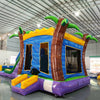 Side angle view of Goombay Splash Dual Lane Commercial Inflatable Combo in a warehouse environment. Shows the full length of the inflatable with dual curved blue slides, palm tree pillars, yellow and blue bounce house area, and splash pool. Other inflatable products and warehouse materials in the background demonstrate its commercial application.