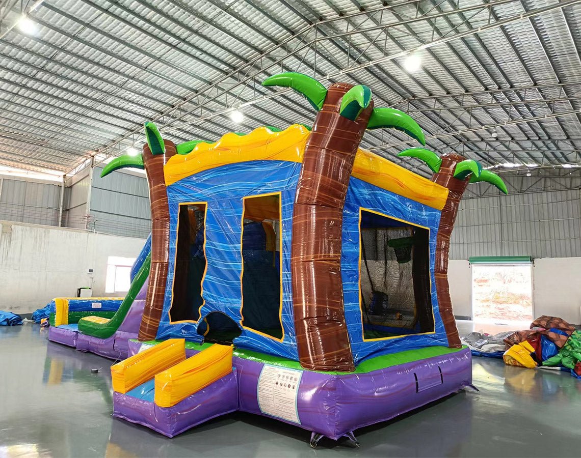 Side angle view of Goombay Splash Dual Lane Commercial Inflatable Combo in a warehouse environment. Shows the full length of the inflatable with dual curved blue slides, palm tree pillars, yellow and blue bounce house area, and splash pool. Other inflatable products and warehouse materials in the background demonstrate its commercial application.