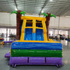 Front view of Goombay Splash Dual Lane Commercial Inflatable Combo (Wet/Dry) set up in a large indoor warehouse. The inflatable features dual curved blue slides flanked by inflatable palm trees with green leaves and brown trunks. A yellow canopy connects the palm trees at the top. The base has a colorful design with blue, green, and purple sections, including a splash pool area. Various inflatable products and warehouse equipment are visible in the background, showcasing its size and commercial setting.