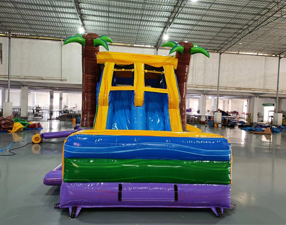 Front view of Goombay Splash Dual Lane Commercial Inflatable Combo (Wet/Dry) set up in a large indoor warehouse. The inflatable features dual curved blue slides flanked by inflatable palm trees with green leaves and brown trunks. A yellow canopy connects the palm trees at the top. The base has a colorful design with blue, green, and purple sections, including a splash pool area. Various inflatable products and warehouse equipment are visible in the background, showcasing its size and commercial setting.