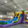 Wide-angle view of Goombay Splash Dual Lane Commercial Inflatable Combo in a spacious warehouse. The inflatable features dual curved blue slides, palm tree pillars, a yellow and blue bounce house structure, and a large splash pool area. Various other inflatable products and warehouse equipment visible, showcasing its size and commercial setting.